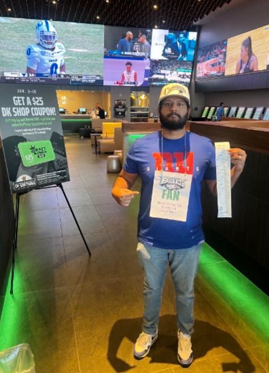 Joel at a DraftKings sportsbook in a Giants shirt, Cowboys hat, and an Eagles fan sign reading he stinks at fantasy football